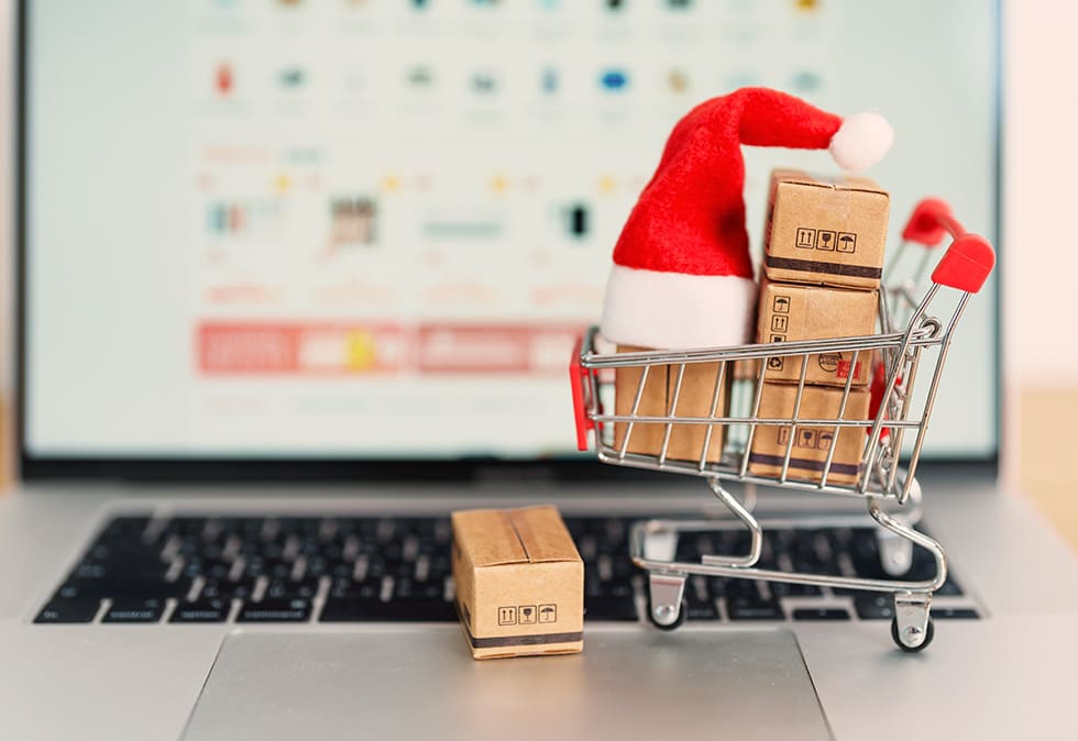a miniature shopping cart packed with parcels, wearing a Santa hat ,with a seasonal marketing plan on the laptop in the background.