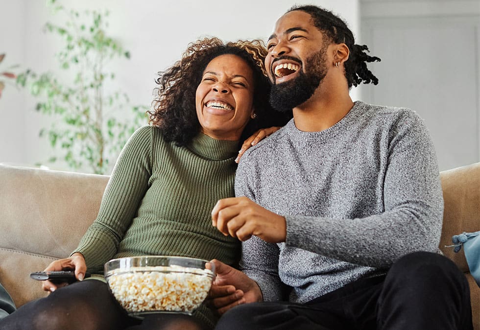 a man and woman sitting on a couch eating popcorn and laughing at fun holiday ads